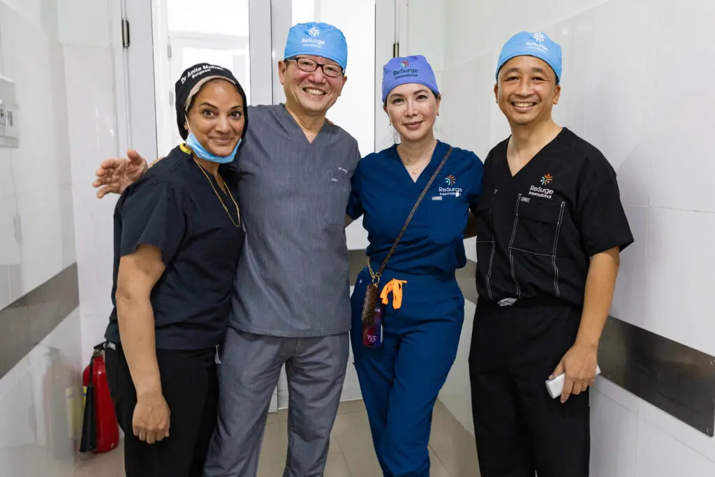 Four healthcare professionals in scrubs and surgical caps pose together in a hospital hallway as a supportive community dedicated to surgery.
