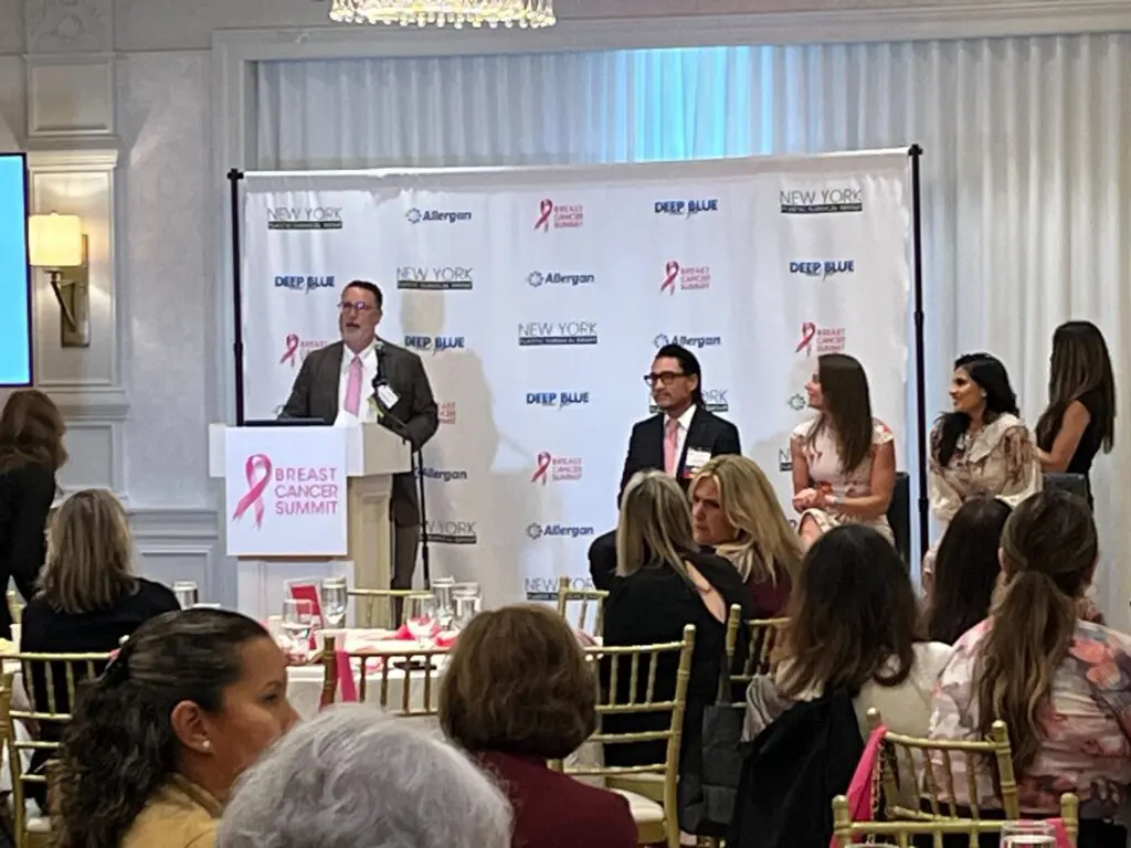 A man speaks at a Breast Cancer Summit podium in front of a banner with corporate logos, while an audience sits at tables in the foreground.