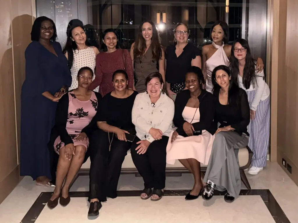 Thirteen Pioneering Women in Reconstructive Surgery (PWRS) pose together indoors, smiling at the camera. Eleven stand while two sit on a bench in an elegant space with wooden walls and marble flooring.