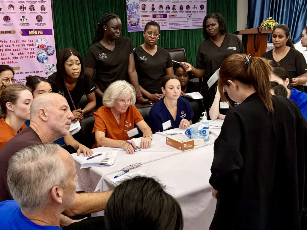 A diverse group of healthcare professionals in scrubs gathers around a table, listening to a woman speak—highlighting leadership like Pioneering Women in Reconstructive Surgery (PWRS). Medical posters and a green curtain are visible behind them.