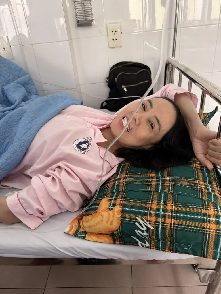 A patient in a pink uniform lies in a hospital bed, receiving support through a nasal cannula.