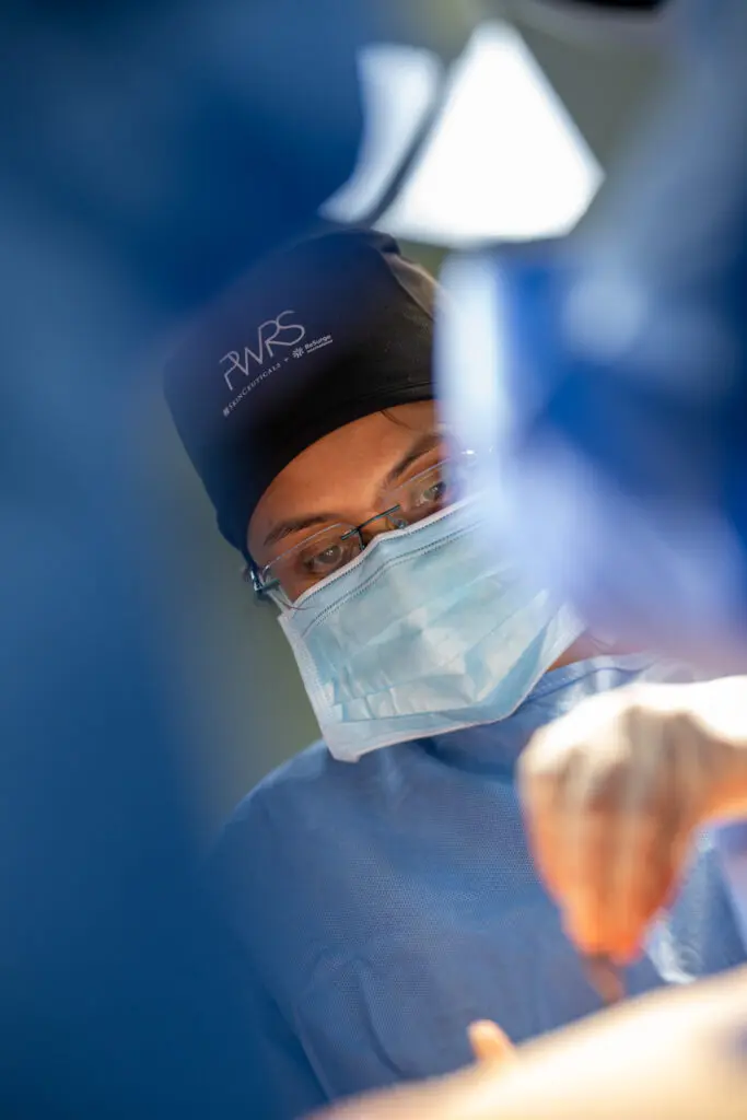 A surgeon wearing a blue face mask and a branded Pioneers in Women’s Reconstructive Surgery cap performs a procedure.
