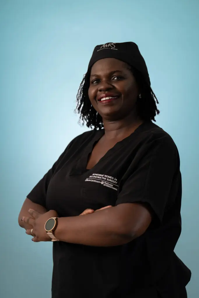 A smiling woman, one of the Pioneering Women in Reconstructive Surgery, stands with her arms crossed against a light blue background while wearing black surgical scrubs and a matching cap.