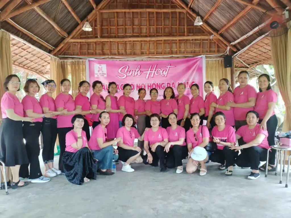A large group of women in pink shirts pose for a breast cancer awareness photo in front of a banner that reads “Sinh Hoạt Câu Lạc Bộ Nơ Hồng Đà Nẵng.”