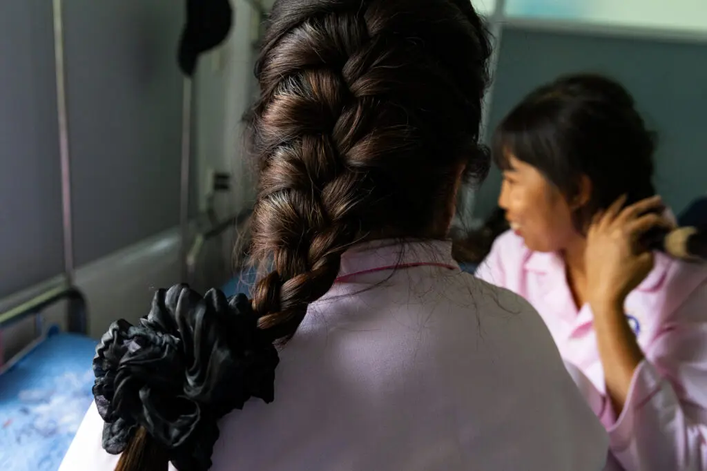 A person with a dark braid secured by a black scrunchie sits in a hospital room wearing pink while another person brushes their hair, reflecting a moment of breast cancer awareness and care.