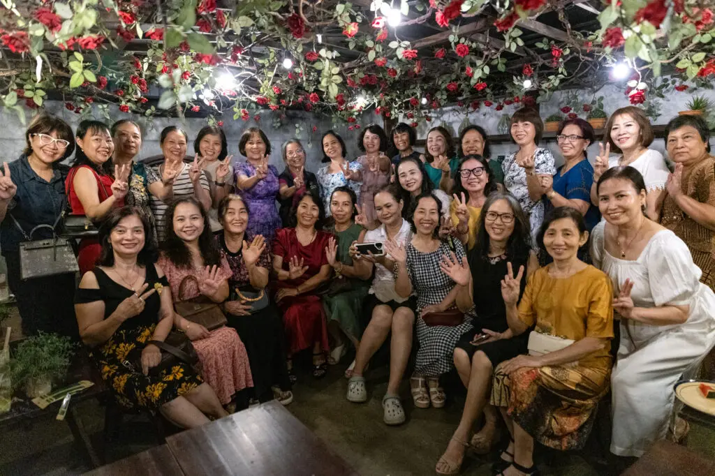 A large group of women in pink posing and waving for a cancer awareness photo under a ceiling covered in red roses and string lights.