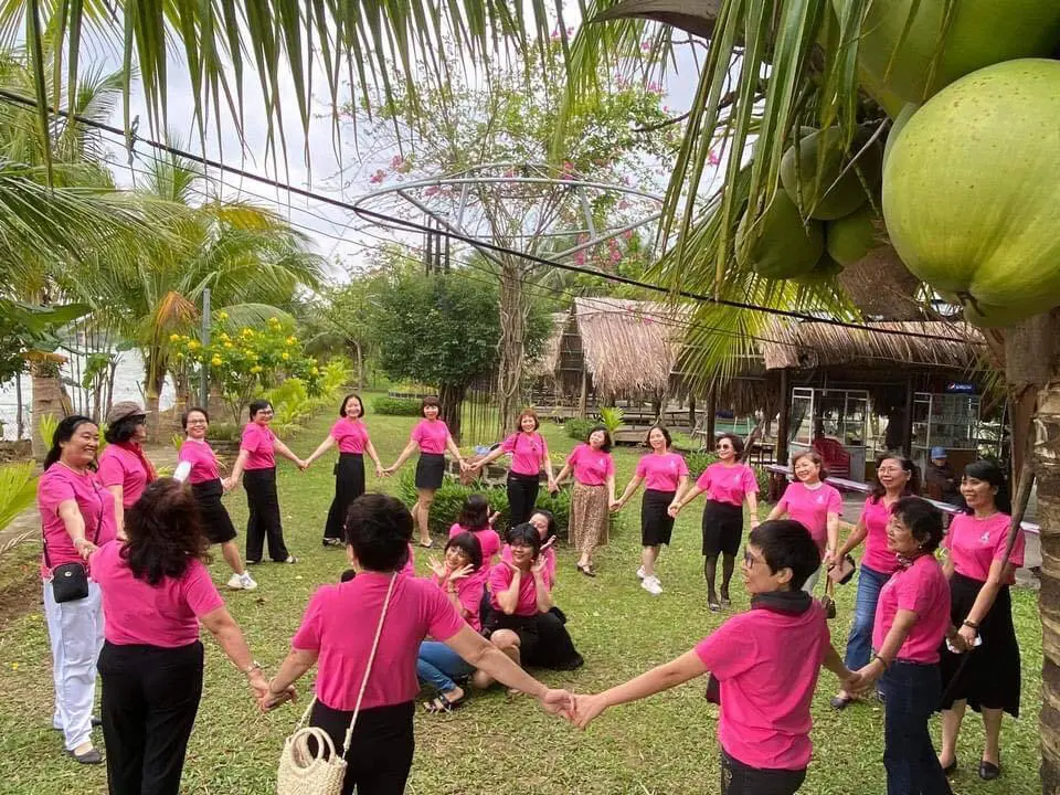 A large group of women wearing matching pink t-shirts for breast cancer awareness hold hands in a circle on a grassy lawn.
