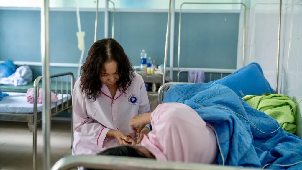A healthcare worker in a white uniform tends to a patient recovering from breast reconstruction after cancer while resting in a hospital bed.