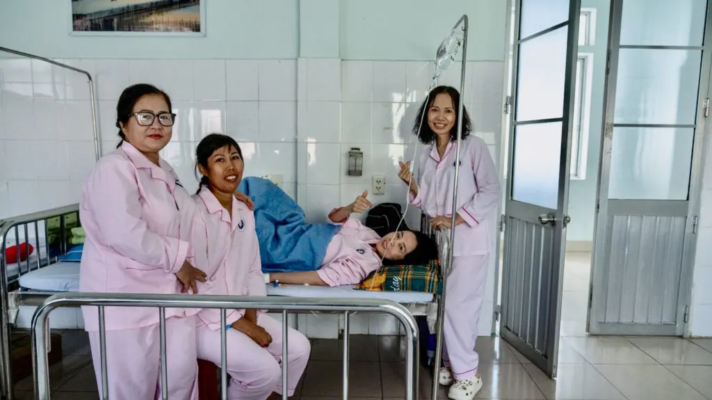 Three women in matching pink hospital pajamas stand around a hospital bed where a fourth woman, also in pink, recovers after breast reconstruction surgery while connected to an IV drip.