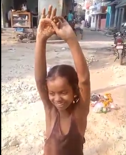 A young girl smiling with her hands raised above her head while holding a small object in a village street.