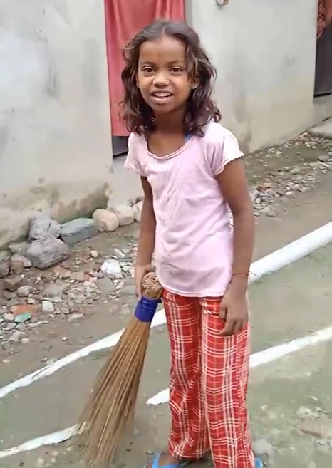 A young girl in a pink shirt and plaid pants holds a traditional straw broom outdoors.