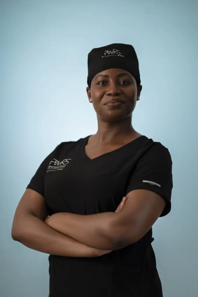 A Pioneering Woman in Reconstructive Surgery stands with arms crossed, wearing black scrubs and a scrub cap against a light blue background.