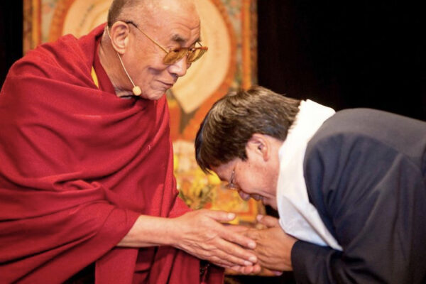 A man in a red robe warmly clasps the hands of another man who is bowing respectfully, both exchanging a gesture of greeting or blessing in front of a colorful background.