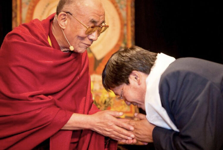 A man in a red robe warmly clasps the hands of another man who is bowing respectfully, both exchanging a gesture of greeting or blessing in front of a colorful background.