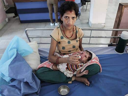 A woman sits cross-legged on a hospital bed, feeding a baby with a bottle in what appears to be Aastha's Story. The baby has visible injuries or burns, while medical supplies and a bowl rest nearby on the bed.
