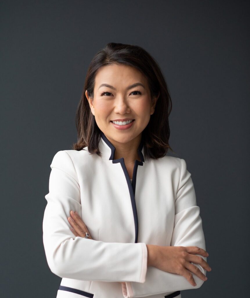 A woman with shoulder-length dark hair, wearing a white blazer with dark trim, stands against a dark background, smiling with arms crossed.