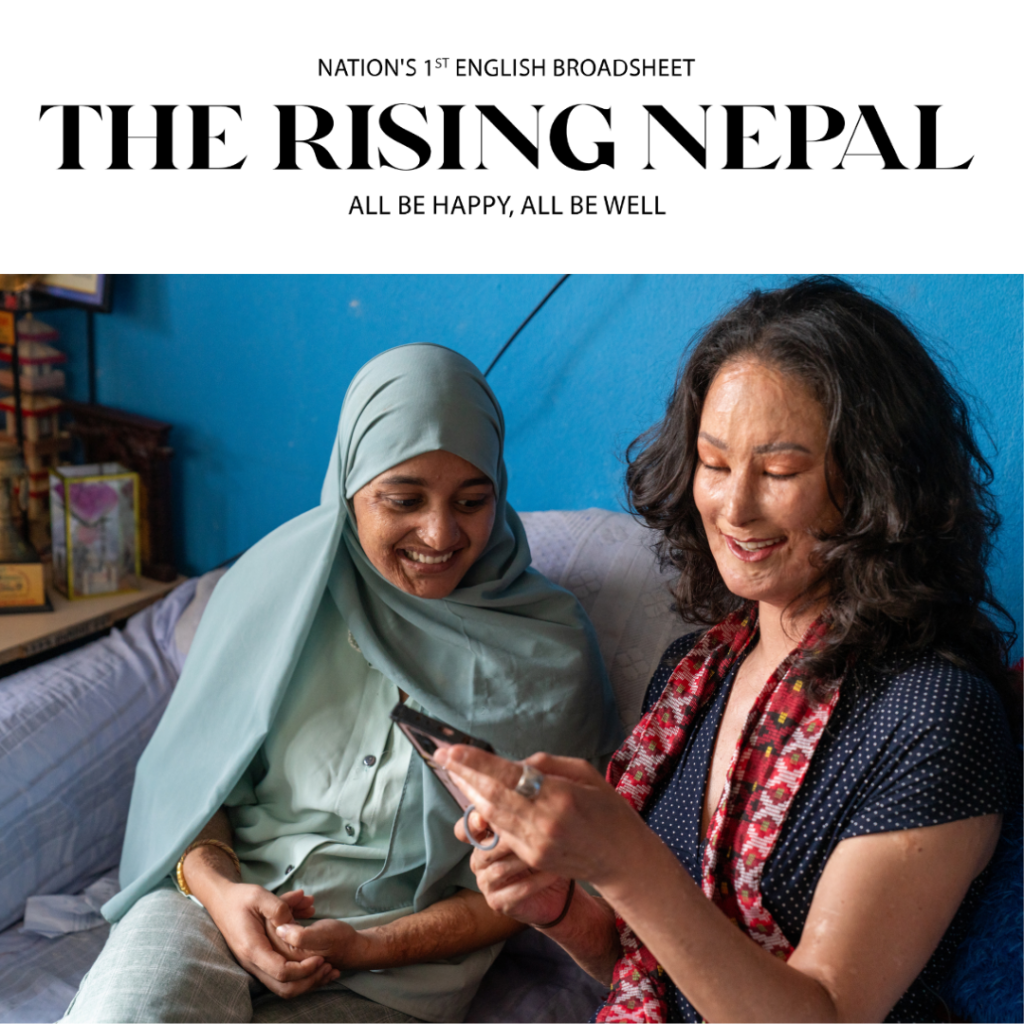 Two women, inspired by Natalia Ponce de León and Muskan Khatun, sit together on a couch, smiling at a smartphone. One wears a light blue hijab; the other has long hair and a floral dress. Behind them is a bright blue wall with shelves and decorations.
