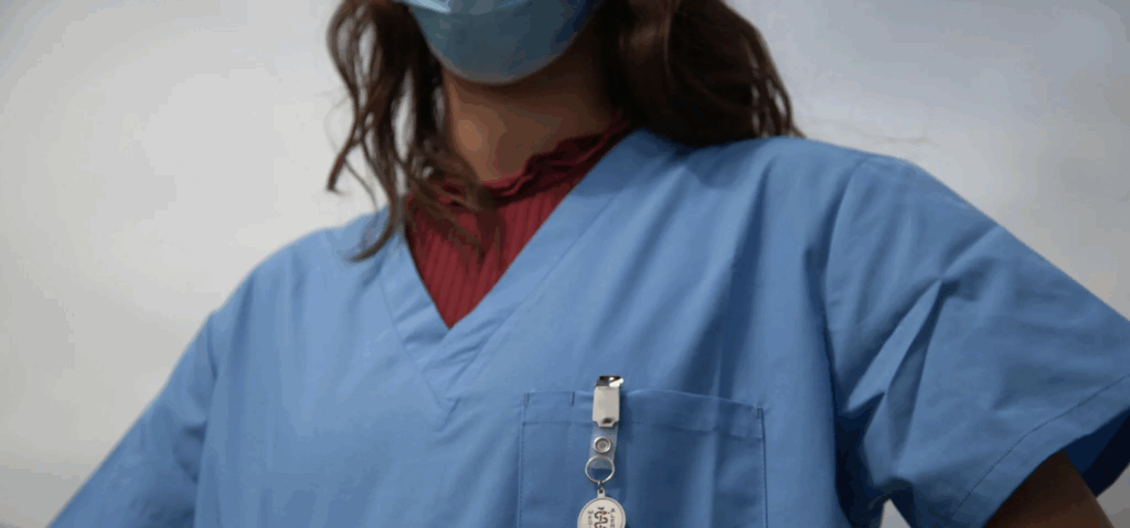 A member of Women in Reconstructive Surgery stands outdoors in blue medical scrubs, a red shirt underneath, and a face mask, with a badge on a retractable lanyard. The sky stretches across the background.
