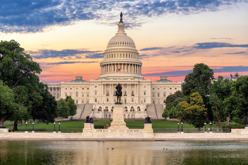 The U.S. Capitol building at sunset, with a colorful sky in the background, trees on both sides, a statue in the foreground, and its reflection shimmering in a pool—symbolizing progress and unity in global health.