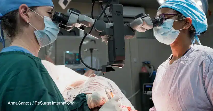 Two surgeons wearing scrubs, masks, and surgical caps are performing breast cancer surgery using a large microscope in an operating room. One surgeon holds surgical instruments while they both look through the microscope.