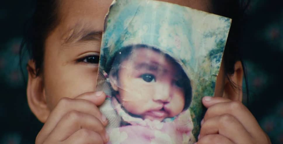 A child holds a worn photo of a baby in front of their face, aligning the image so one eye is visible next to the photo. The dark, blurred background adds depth—highlighting why you should consider donating to ReSurge.