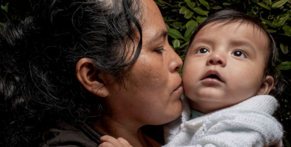 A woman with dark hair kisses a baby on the cheek. The baby, in a white sweater, looks upward with a calm expression. Green leaves are visible in the blurred background, reminding us to cherish every moment—Donate to ReSurge and help transform lives.