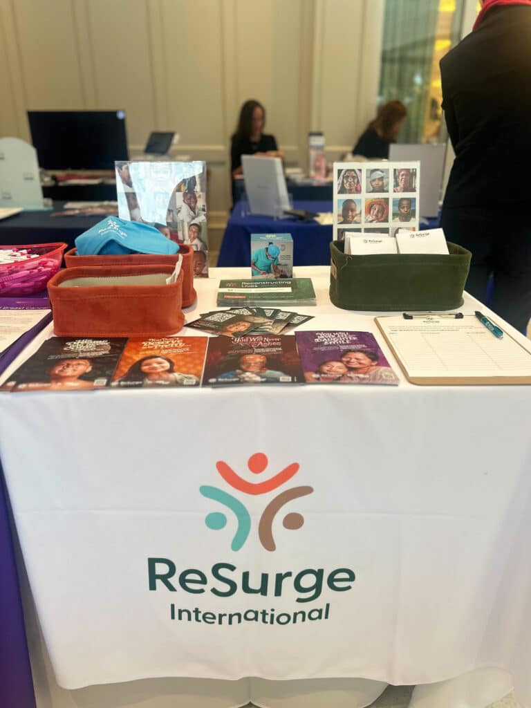 A display table for ReSurge International features informational brochures on breast reconstruction, photos, business cards, and a clipboard, all arranged on a white tablecloth with the organization's logo. People are visible in the background.