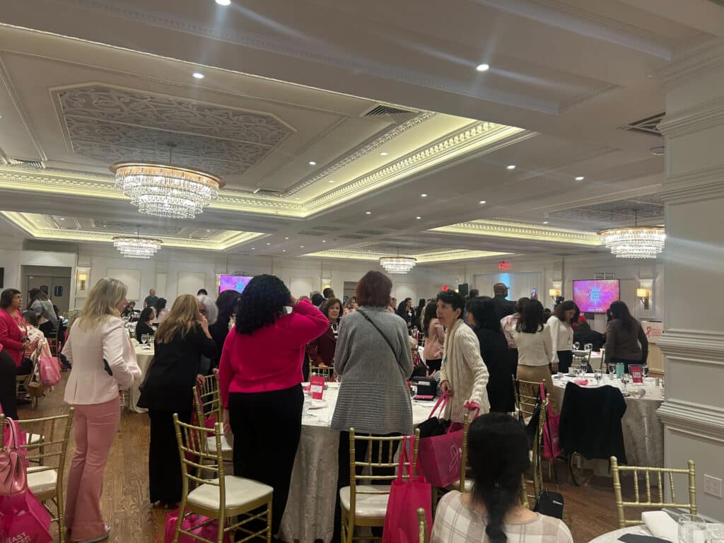A large group of people, mostly women, gather around decorated tables in an elegant ballroom with chandeliers and pink-themed décor, attending a formal event focused on breast cancer care and awareness.
