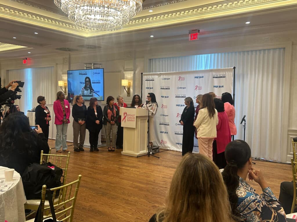 A group of women stand on a stage near a podium at an indoor event focused on breast cancer care. One woman speaks at the podium while others stand beside her. Attendees sit at tables under a large chandelier.