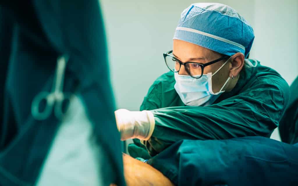 A focused surgeon wearing a mask, glasses, and surgical cap performs an operation related to breast reconstruction, reaching toward a patient. Surgical instruments and a medical team member are partially visible in the foreground.
