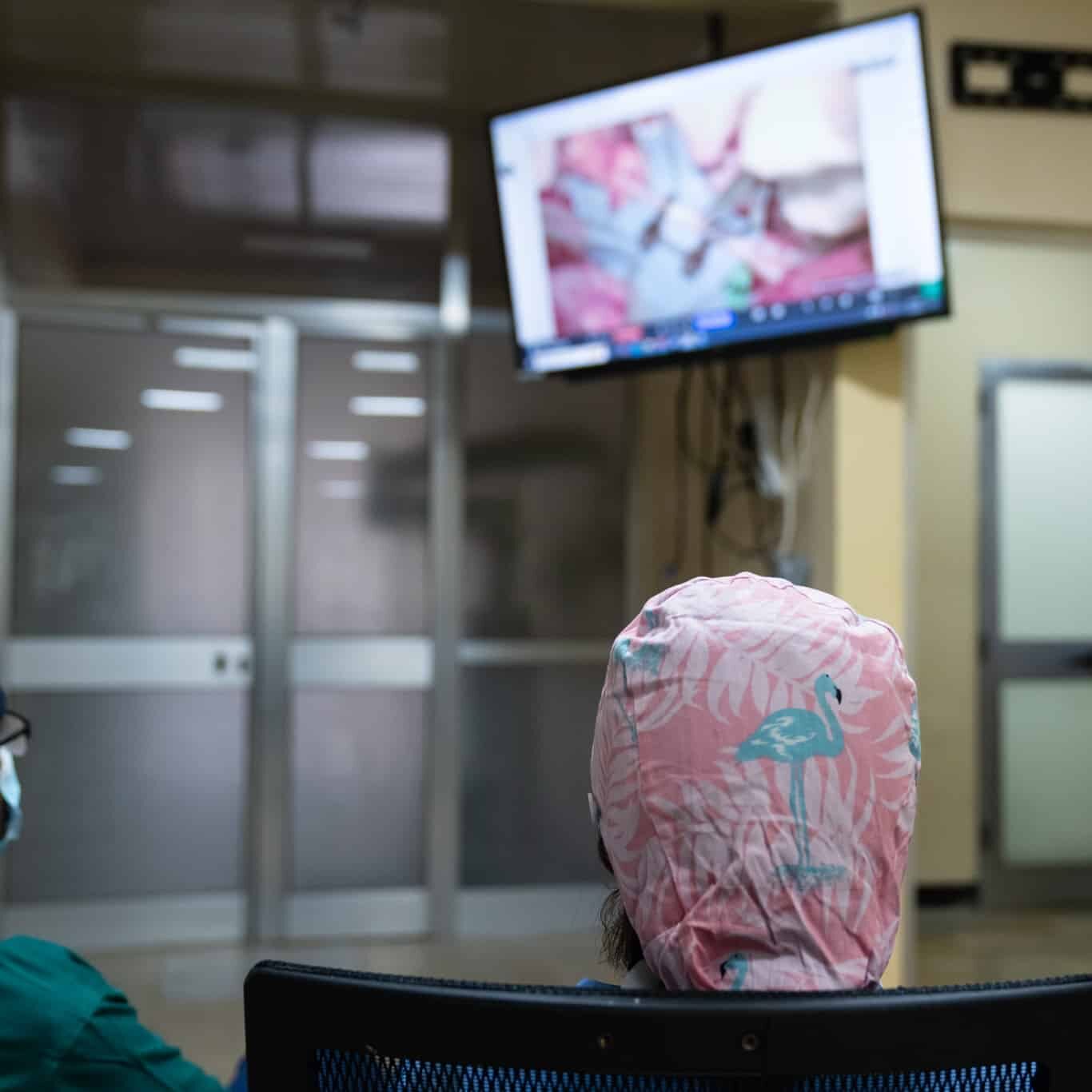 Three medical professionals in scrubs and caps watch a surgical procedure on a wall-mounted monitor in a hospital room, echoing the collaborative spirit seen at events like the World Health Assembly. The scene is viewed from behind the seated viewers.
