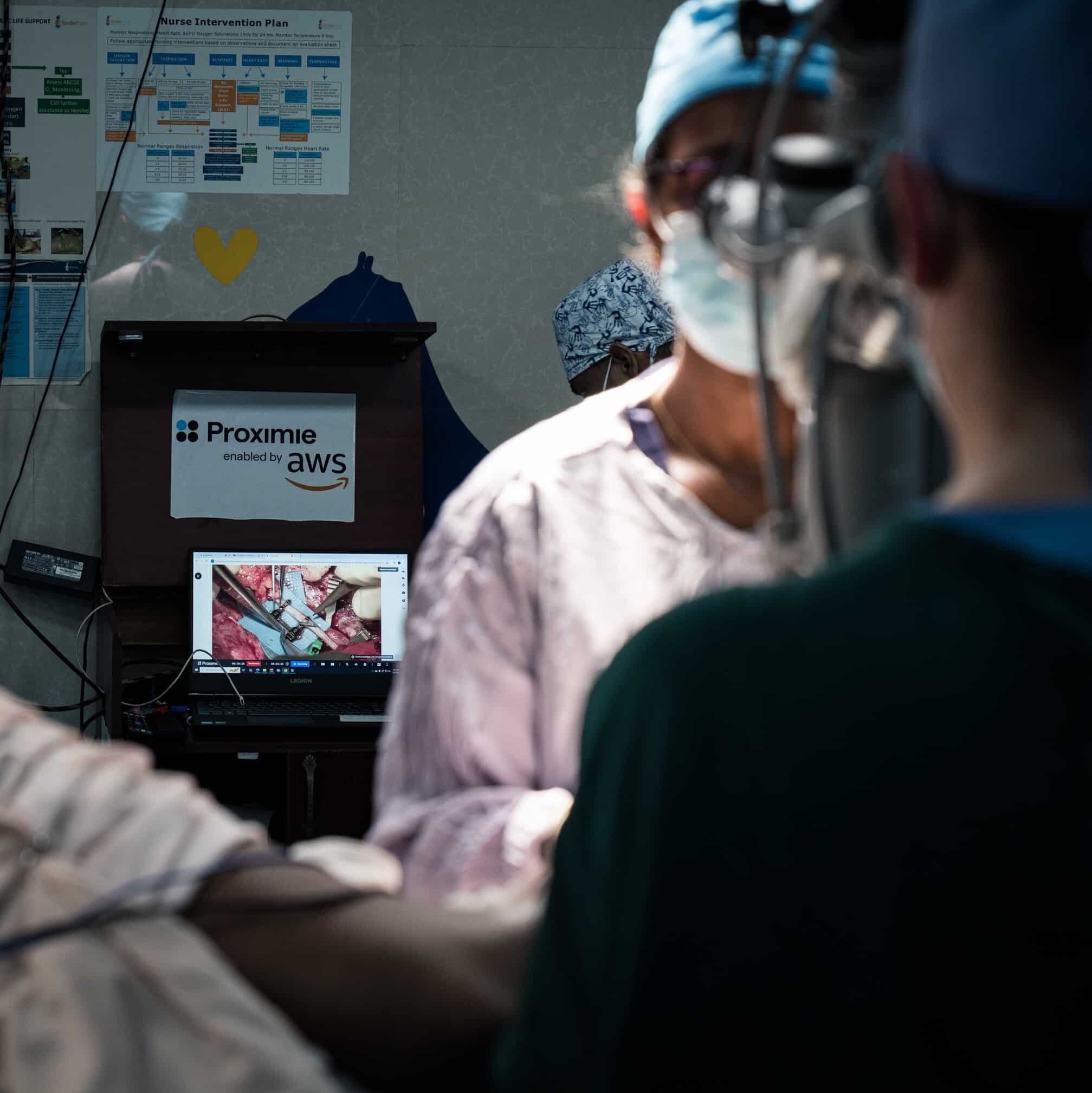 Two medical professionals in surgical attire perform a procedure, with a laptop displaying a live surgical video in the background. A sign on the laptop stand shows "Proximie enabled by AWS," supporting innovation discussed at WHA79, the World Health Assembly.