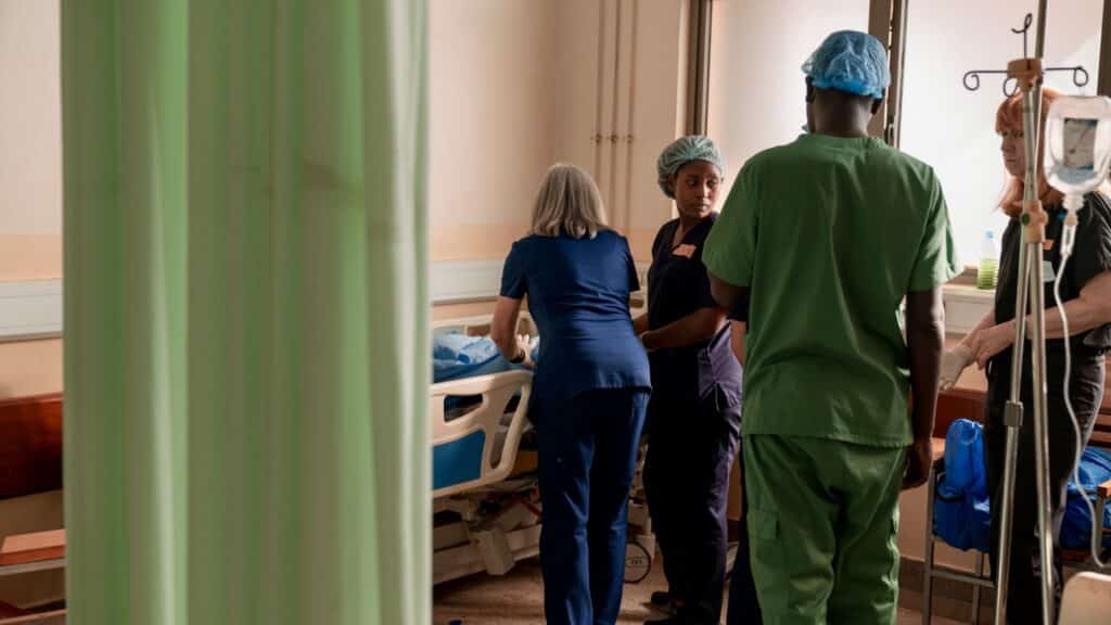 A group of healthcare workers, dressed in scrubs and surgical caps, stand around a hospital bed in a patient room, preparing for thumb reconstruction surgery on a lost thumb. Medical equipment and IV stands are visible—part of ReSurge’s vital work.