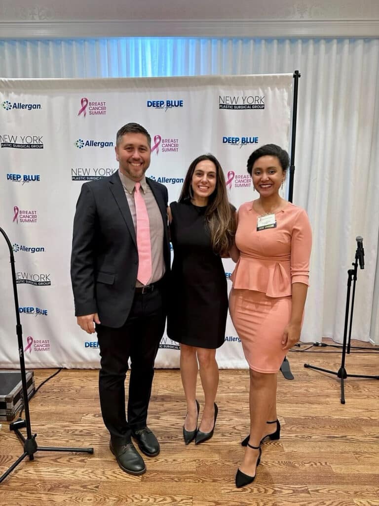 Three people stand smiling before a step-and-repeat banner at a breast cancer event focused on breast cancer care. Two women wear black and pink dresses, while the man sports a suit with a pink tie amid sponsor logos in the background.