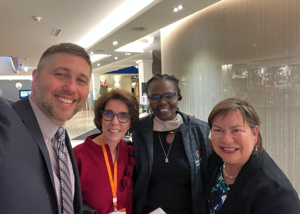 Four adults, three women and one man, are smiling and posing together indoors in a well-lit room, dressed in business or professional attire at the WHA79 World Health Assembly.