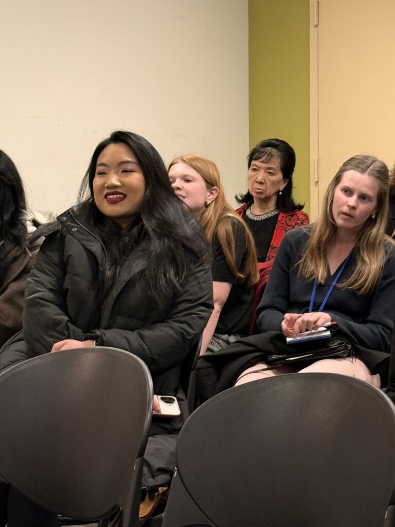 Four women are seated indoors, perhaps attending a session about breast cancer care. One woman in a black coat is smiling, while the others appear attentive. Empty chairs sit in the foreground; the wall behind them is light yellow and green.