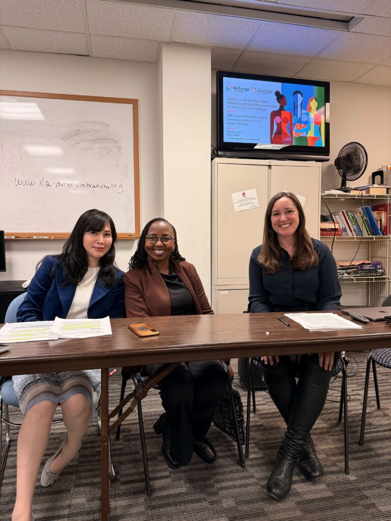 Three women are seated at a table with papers in front of them, smiling at the camera. Behind them are bookshelves, a whiteboard, and a TV screen displaying a colorful graphic about breast cancer care.