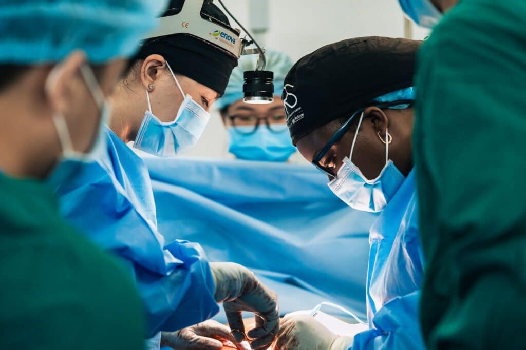 A team of surgeons wearing masks, scrubs, and surgical caps operates in a brightly lit operating room, focused on a patient beneath a blue sterile drape. One surgeon wears a headlamp for extra illumination.
