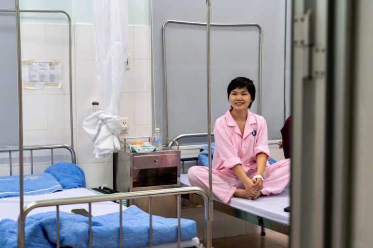 A woman in pink pajamas sits cross-legged and smiling on a hospital bed in a hospital room with another empty bed, blue bedding, and medical supplies nearby.