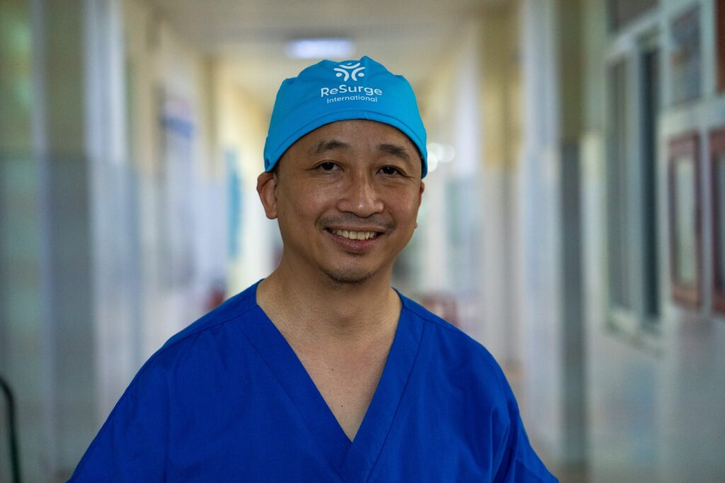 A smiling person in blue medical scrubs and a blue cap with "ReSurge International" logo stands in a hospital hallway. The background is softly blurred, highlighting the individual.