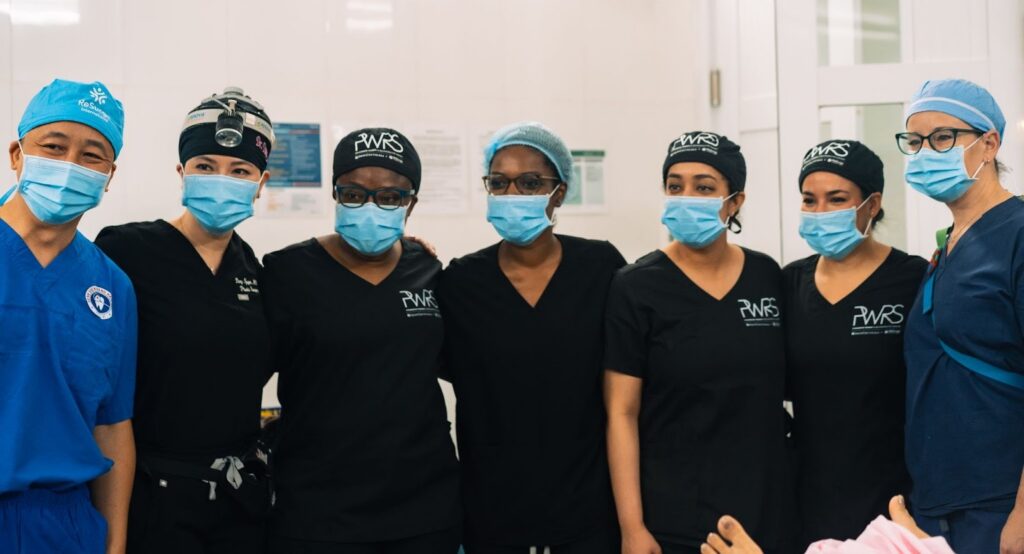A group of seven healthcare professionals in scrubs and face masks pose together in a bright, clean medical room, showcasing their dedication to breast cancer care. Some wear surgical caps and headlamps, highlighting their expertise in this specialized field.