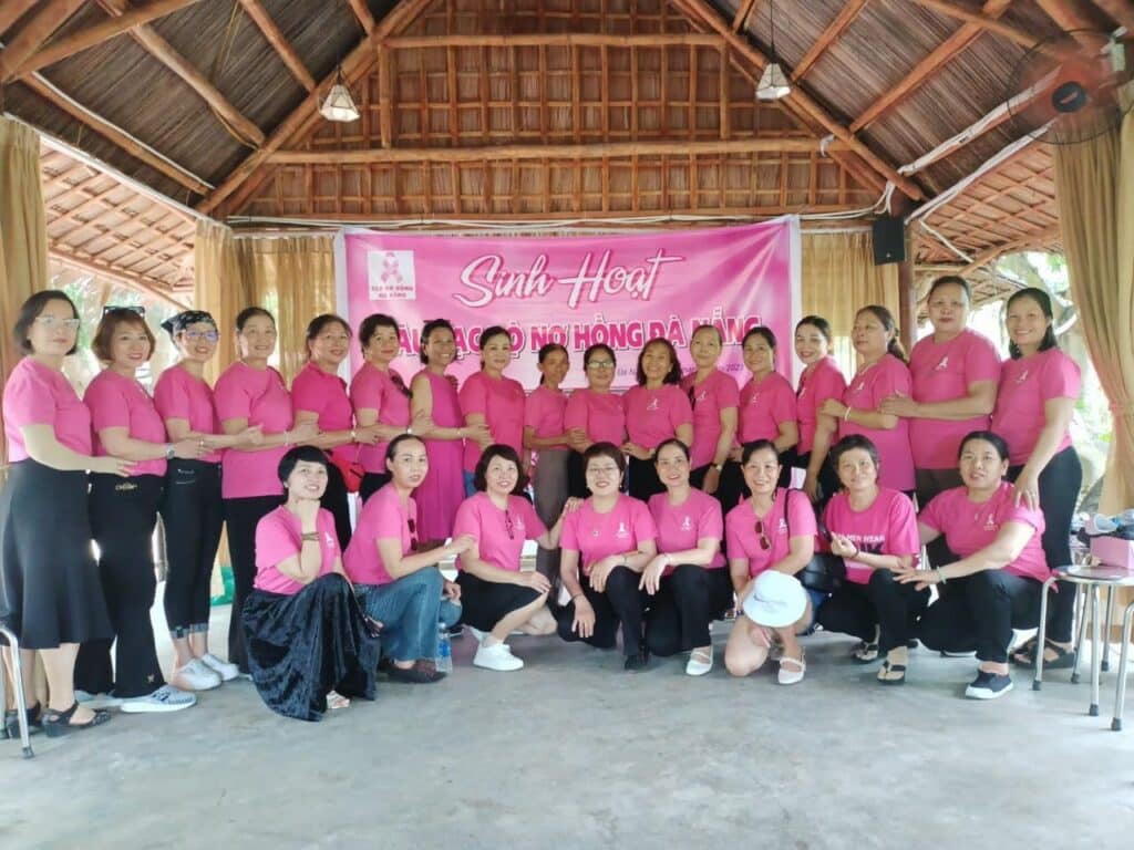 A group of women wearing matching pink shirts pose together indoors under a thatched roof, standing in front of a pink banner with Vietnamese text. Smiling and linking arms, they celebrate unity and support for breast cancer care.