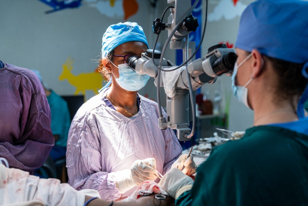 Two surgeons in scrubs and masks perform breast reconstruction surgery using a microscope in a brightly lit operating room with animal decorations on the wall in the background.