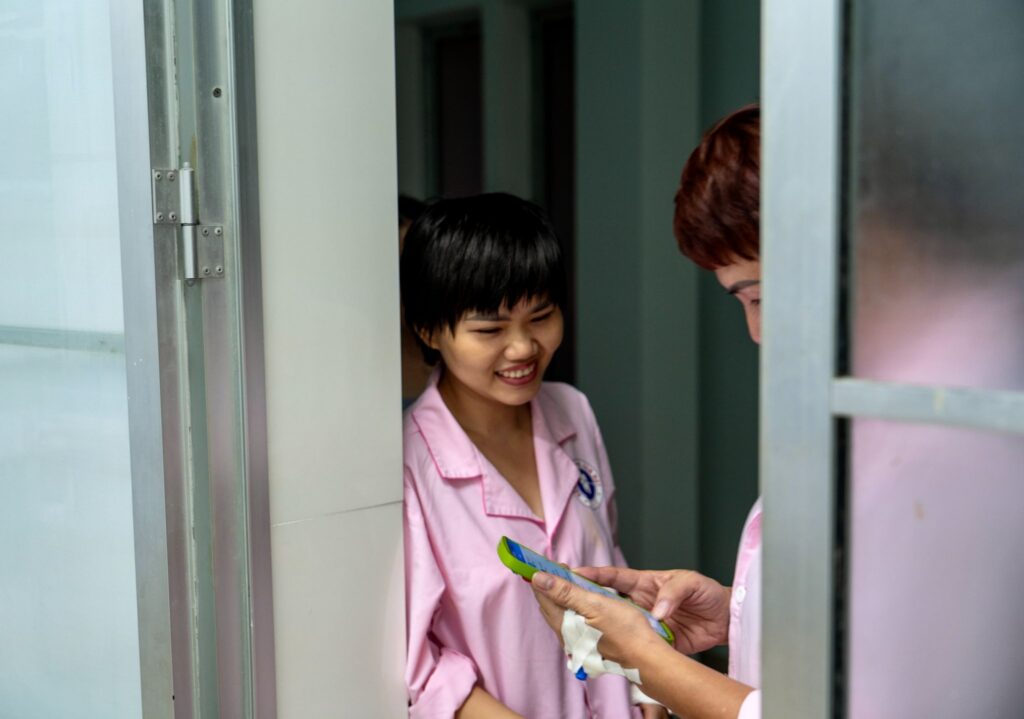 Two people in pink uniforms stand by a doorway. One smiles while the other, partially visible, holds a smartphone and tissue—capturing a moment of dedication often seen in breast cancer care within medical or institutional environments.