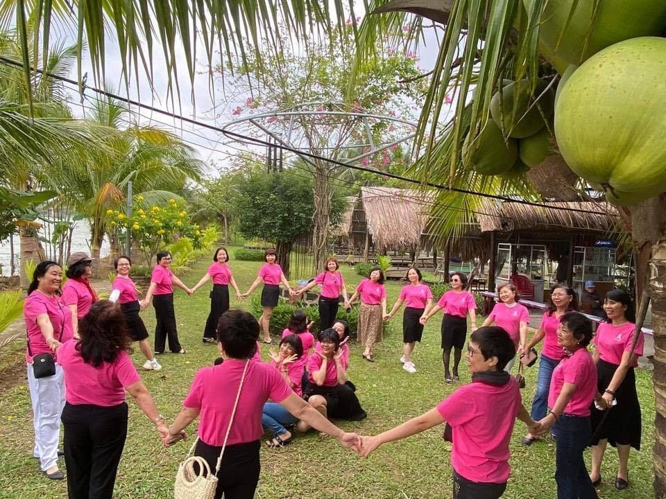 A group of women wearing matching pink shirts stand in a large circle holding hands outdoors on a grassy area, surrounded by trees and huts, participating in a team-building activity.