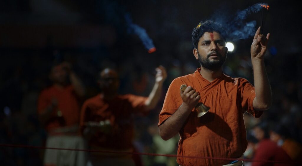 A man in orange traditional attire holds a bell and incense, performing a ritual—a scene reminiscent of moments captured in a reconstructive surgery Nepal documentary film. Two other people in similar attire are blurred in the background, also participating in the ceremony, as smoke rises from the incense.