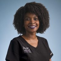 A woman with curly dark hair and purple lipstick smiles at the camera. She is wearing a black scrub top with white lettering, representing Women in Reconstructive Surgery. The background is a plain light blue.