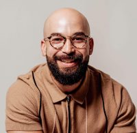 A smiling man with a bald head, beard, and glasses—Dr. Félix Chinea—wearing a brown collared shirt, poses against a plain light background.