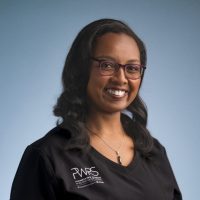 A woman with long dark hair, wearing glasses and a black top with a "PWRS" logo smiles at the camera against a plain blue background. She represents Women in Reconstructive Surgery and wears a silver necklace.