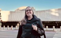 A woman with glasses and long hair, wearing a dark coat, smiles while standing on a city street with a modern, dome-shaped building in the background under a clear blue sky.